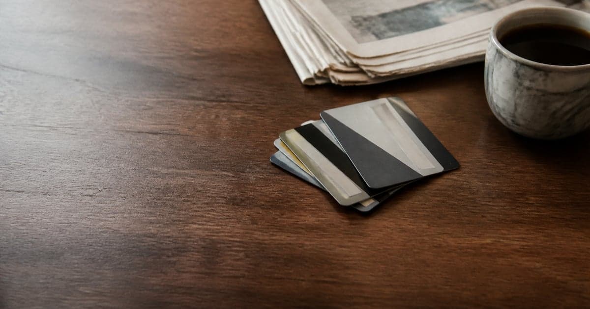 several credit cards fanned face-down on a dark walnut table beside a folded newspaper and a cup of black coffee