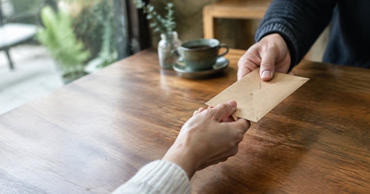two hands passing a small brown paper envelope across a wooden café table